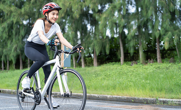 Mulher asiática com blusa branca e calça preta andando de bicicleta com capacete.