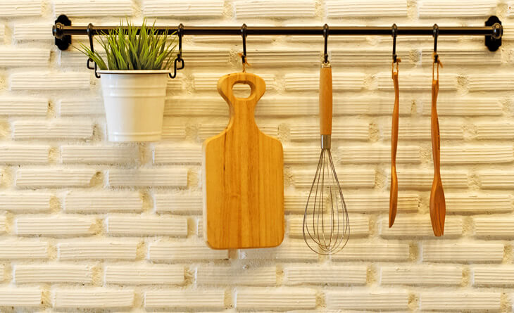 TÃ¡bua de madeira, batedor de claras e colheres pendurados em ganchos na parede da cozinha.