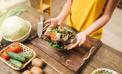 Mãos segurando um prato de salada em cima de uma bancada com legumes e verduras.