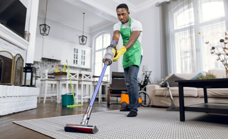 homem de avental verde tirando pó do tapete com o aspirador.