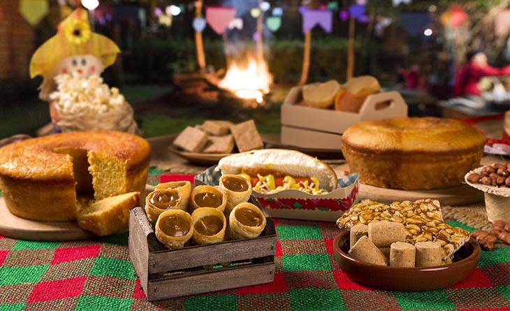 Mesa de festa junina com toalha xadrez, bolo de milho, hot dog, paçoca, pé de moleque e canudinho de doce de leite.