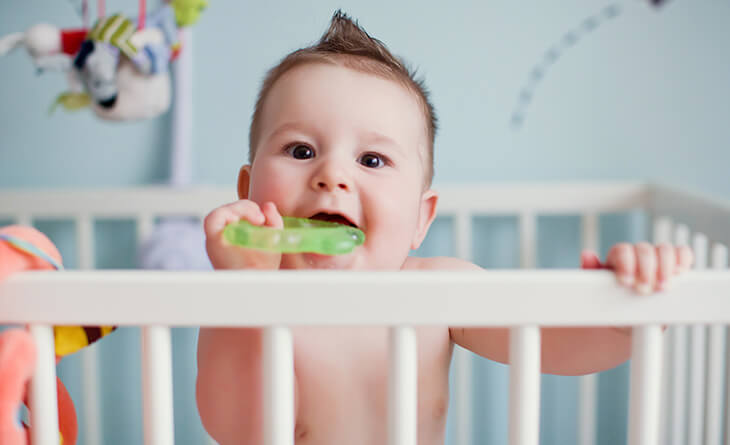 Bebê com topete segurando um mordedor de plástico verde dentro do berço.
