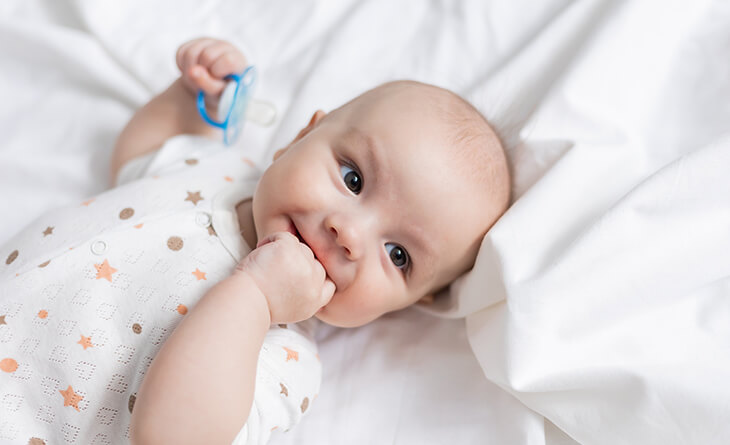 Bebê usando roupa branca deitado na cama com lençol branco, segurando uma chupeta azul.