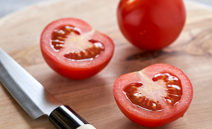 Um tomate cortado na metade e o outro inteiro em cima de uma tábua de madeira e ao lado de uma faca.