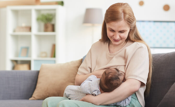 Mulher amamentando o bebê no sofá da sala.