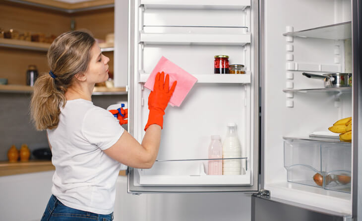 Mulher com luva laranja limpando a geladeira com uma flanela rosa.