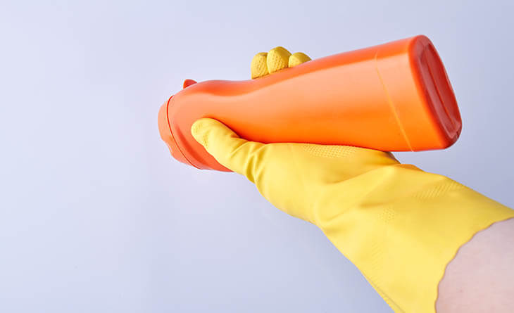 Mulher de luva amarela segurando um produto de limpeza com embalagem laranja.