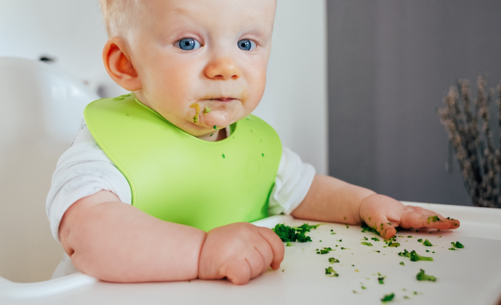 Bebê comendo legume na cadeirinha de alimentação.