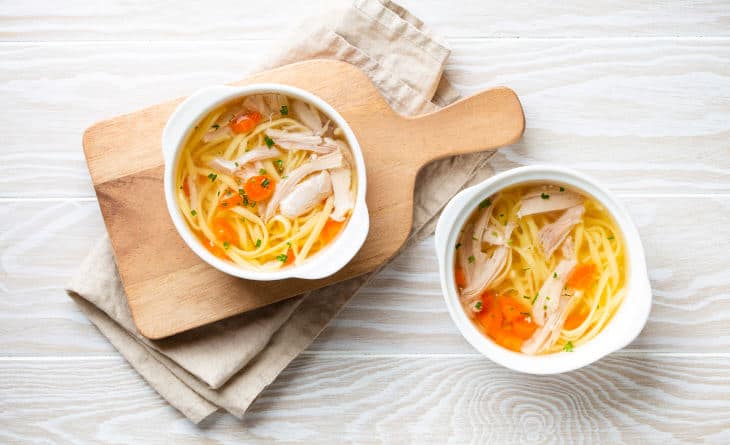 Sopa de galinha com legumes em tigelas brancas sobre mesa de madeira.