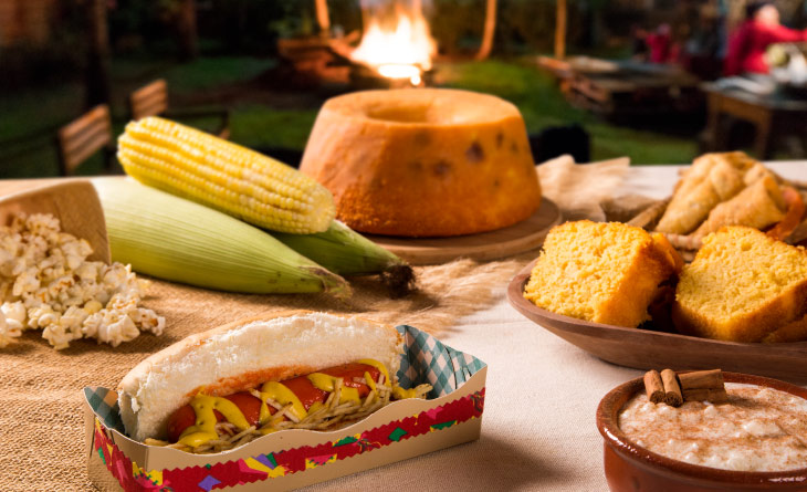 Mesa com comidas de festa junina: milho, bolo de fubá com goiabada, cachorro quente e pipoca.