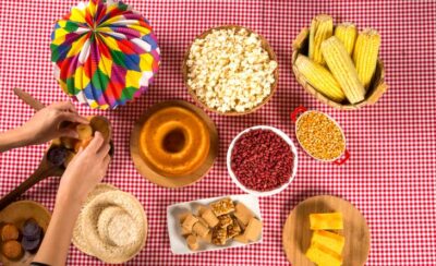Mesa com toalha quadriculada vermelha e branca com comidas de festa junina.