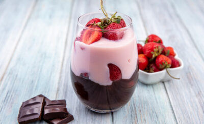 Bombom de morango num copo de vidro ao lado de morangos frescos e pedaços de chocolate.