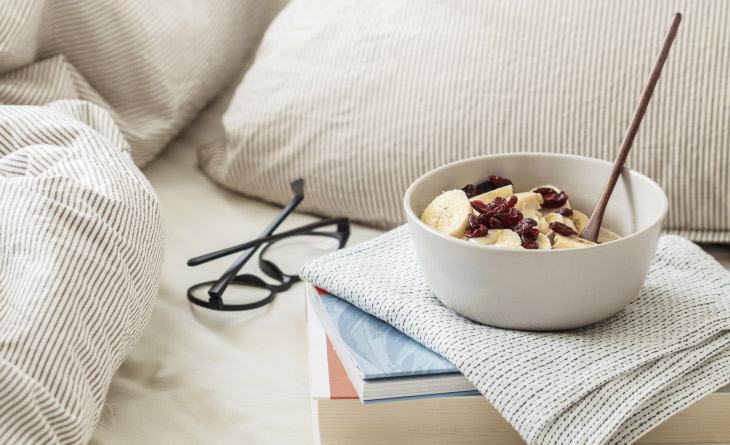 cama ao fundo com cumbuca de banana com aveia sobre móvel a frente representando alimentos que ajudam a dormir