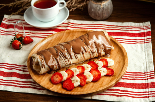 Prato com panqueca de chocolate ao lado de morangos e bananas picadas, próximo a uma xícara de chá.