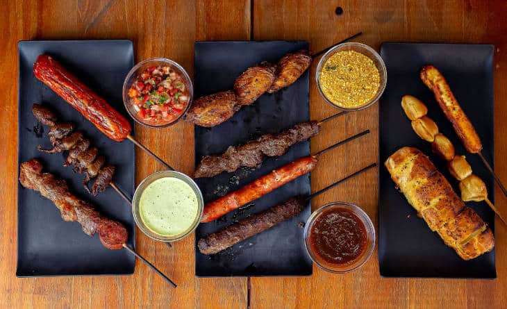 Espetos de churrasco em dois pratos retangulares pretos e potes com vinagrete.
