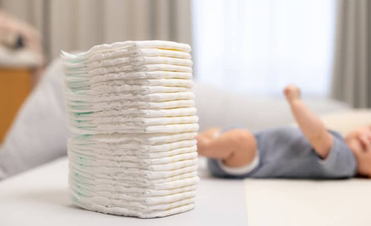 Pilha de fraldas descartáveis e bebê ao fundo.