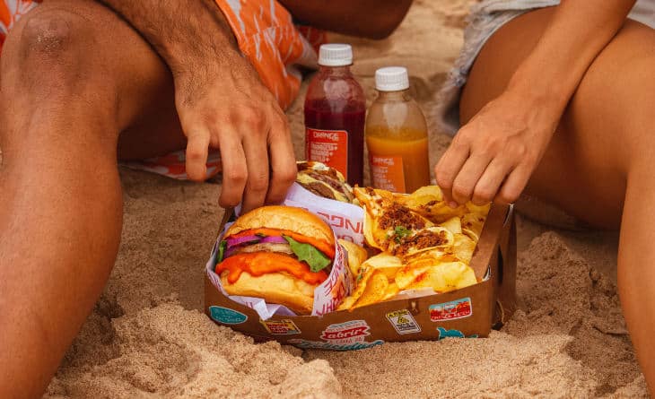 Combo de lanche e batata, perto de sucos, na areia da praia.